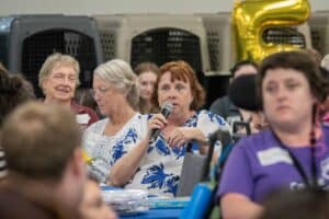 A woman with reddish-brown hair and a floral blue and white blouse speaks into a microphone at a gathering, while two older women listen beside her. In the background, people are seated in a large hall, and a gold balloon shaped like the letter 'E' is visible.