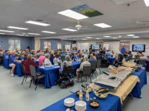 A large gathering in a community hall featuring several tables covered with blue tablecloths, where attendees are enjoying food and watching presentations on screens. The room is filled with people, including some in wheelchairs, and there are trays of food in the foreground.