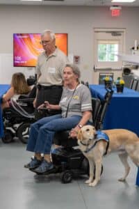 An older man with glasses stands beside a woman in a wheelchair, who is holding a joystick control. The woman has gray hair and wears a gray shirt with a logo, while a Labrador retriever in a blue vest sits next to her. In the background, there are tables adorned with blue tablecloths and flower arrangements.