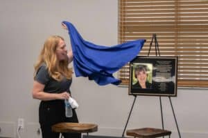 A woman with long, wavy red hair enthusiastically pulls away a blue cloth, revealing a framed portrait and plaque on an easel that honors someone named Margaret S. Ager. The setting appears to be an indoor space with wooden blinds and simple wooden furniture.