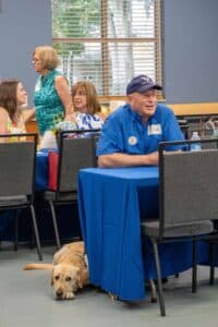 A seated elderly man with a blue shirt and a cap smiles at a gathering, while a yellow Labrador lies quietly on the floor beside him. In the background, several women engage in conversation around blue tablecloths.