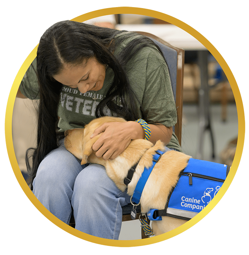 A woman wearing a green t-shirt sits in a chair, gently hugging a yellow Labrador retriever dog that is wearing a blue vest labeled 'Canine Companions.' They share a moment of affection, with the dog resting its head on her lap.
