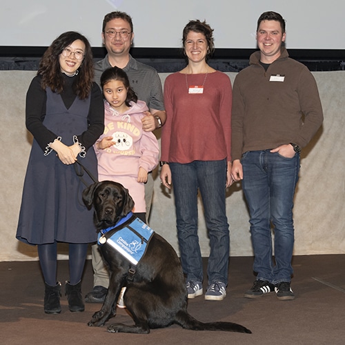 A group of five people, including a woman in a black dress, a man in a grey shirt, a young girl in a pink hoodie, a woman in a red top, and a man in a blue shirt, stand together on stage with a black labrador retriever wearing a service dog vest.