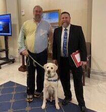 Two men standing alongside a Canine Companions service dog in a formal setting with a blue carpet and decorative walls. One man is wearing a black suit and tie, while the other is dressed in a half-white, half-color shirt.