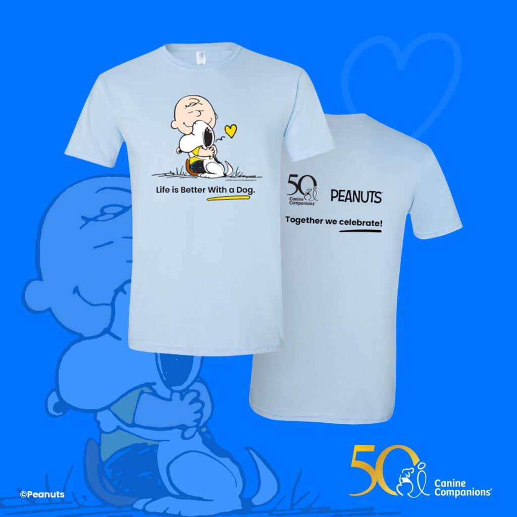 A light blue t-shirt featuring a cartoon character joyfully hugging a dog, with the text 'Life is Better With a Dog.' The back of the shirt displays '50 PEANUTS' and 'Together we celebrate!' alongside the logos for Canine Companions and the 50th anniversary.