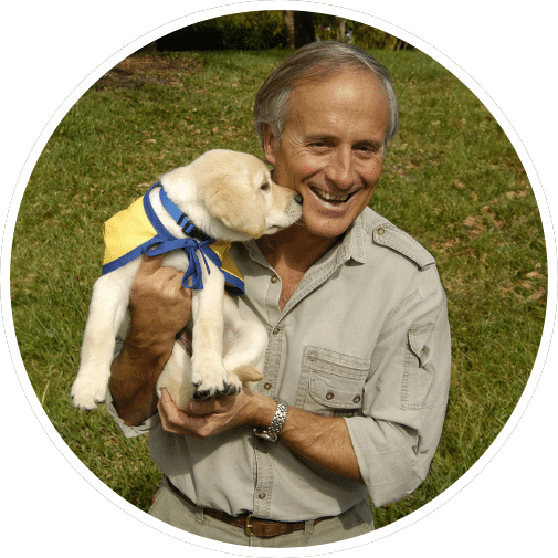 A smiling man in a light-colored shirt holds a small yellow Labrador puppy wearing a blue and yellow harness, as they are surrounded by green grass.