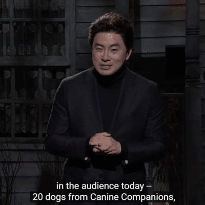 A man in a black turtleneck and blazer is speaking on stage, with a dark, rustic background. He appears to be addressing an audience and mentions that there are 20 dogs from Canine Companions present.