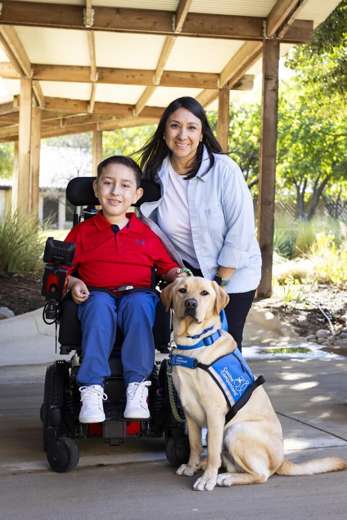 A young boy in a red shirt is seated in a motorized wheelchair, smiling alongside a woman in a denim jacket. Next to them sits a yellow Labrador retriever wearing a blue harness labeled 'Canine Companions.' The scene is set outdoors, under a wooden structure with greenery in the background.