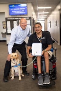 A man in a blue dress shirt poses beside a young man in a black shirt holding a document, who sits in a power wheelchair. A yellow Labrador service dog sits in front of them. In the background, a digital sign displays information about a city council meeting.