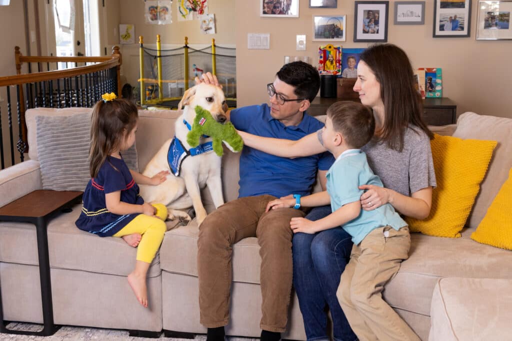 A young family sits on a couch in a living room, interacting with a yellow Labrador retriever wearing a blue service vest. The father is holding a green toy while the mother sits beside him, smiling. A girl in a navy dress with yellow leggings is petting the dog, and a boy in a blue polo shirt is reaching towards the dog as well. Colorful cushions and family photos decorate the background.