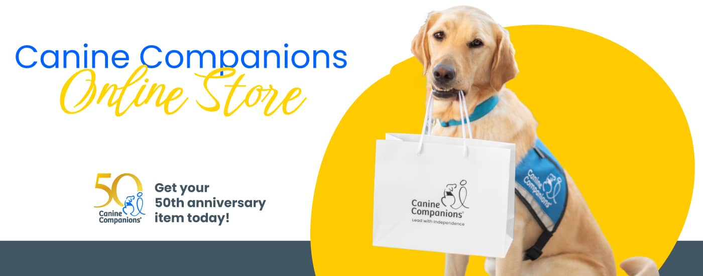 A Labrador Retriever wearing a blue vest with the Canine Companions logo holds a white shopping bag in its mouth, with promotional text for the online store celebrating the 50th anniversary of Canine Companions.