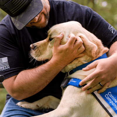 A smiling man in a black t-shirt gently embraces a yellow Labrador Retriever wearing a blue vest with the text "Canine Companions" as they share a moment outdoors in a park setting.