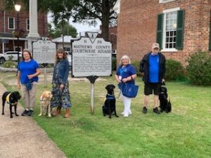 A group of five people with their service dogs, standing in front of a historic sign that reads 'Mathews County Courthouse Square.' They are wearing blue shirts, and the dogs include two black labs and a yellow lab.