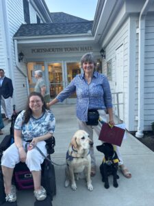 A woman with short gray hair stands next to a seated woman in a wheelchair; the seated woman is holding a leash attached to a yellow Labrador retriever, while a black labrador puppy sits beside them. They are in front of Portsmouth Town Hall.