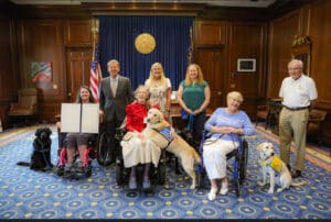 A group photo featuring six people in a formal room with a blue and yellow carpet and wooden walls. Two women in wheelchairs hold a certificate, while three standing women and a man in a suit pose behind them. Two therapy dogs, one black and one yellow, are also present, with one dog sitting on the lap of a woman in a red dress.