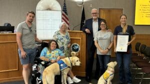 A group of five individuals poses for a photo in a municipal chamber, with two service dogs seated in front. The group includes a man in a grey shirt and shorts, a woman in a blue shirt in a wheelchair, an elderly woman in a floral blouse, a man in a suit, and a woman wearing a grey 'Canine Companions' shirt. One woman holds a proclamation document.