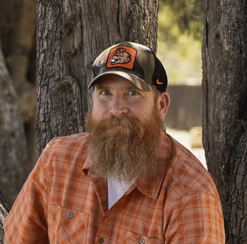 A man with a long beard wearing an orange plaid shirt and a camouflaged cap sits against a tree, smiling at the camera.