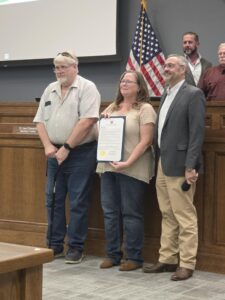 A group of four individuals stands in front of a wooden podium in a meeting room, with an American flag in the background. A woman in the center holds a framed proclamation while smiling, flanked by two men on her sides, one with glasses and a blazer, and another with a cane. In the background, a few other individuals are partially visible.