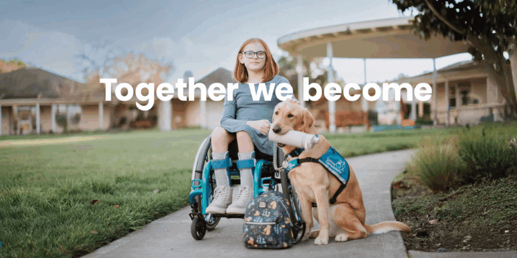 A young girl in a wheelchair with a blue sweater and glasses is smiling next to a service dog, which is holding a toy in its mouth. The background features green grass and a pathway leading to a building. The text 'Together we become' is superimposed in white.