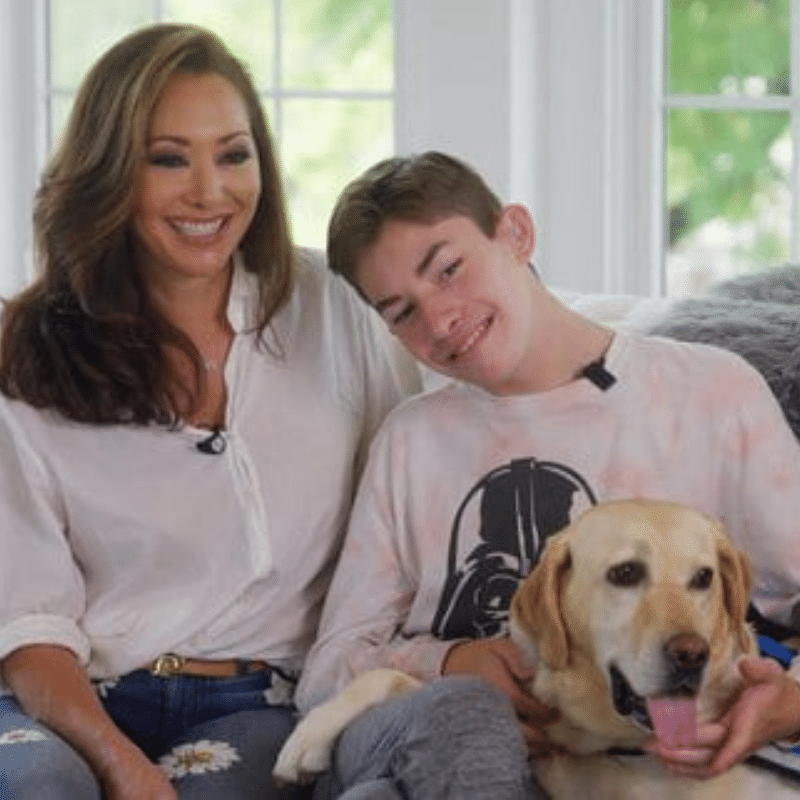 A woman and a teenage boy smile while sitting together on a couch, holding a yellow Labrador Retriever. The woman has long, flowing hair and wears a white shirt, while the boy is dressed in a tie-dye shirt featuring a character. The background shows large windows with natural light.