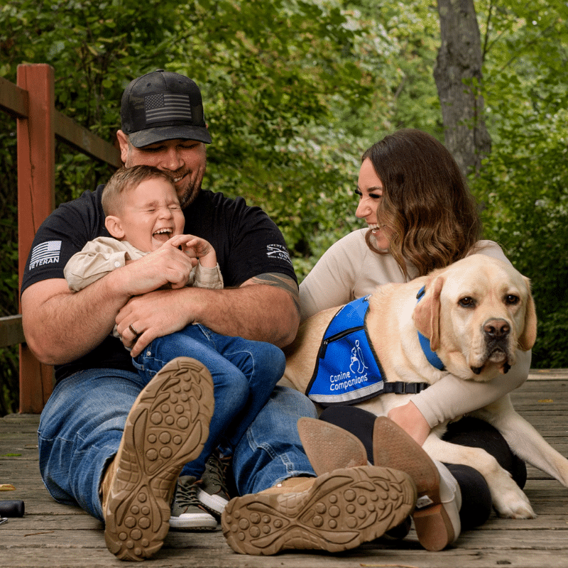 A father and his young son, both laughing, seated on a wooden deck surrounded by greenery. The father, wearing a black cap and a veteran t-shirt, is holding the boy, who is joyfully smiling with his eyes closed. Next to them, a woman is sitting on the deck, smiling at the boy; she has long, wavy hair and is wearing a light sweater. A large, well-groomed yellow Labrador, wearing a blue vest branded "Canine Companions," is resting beside her.