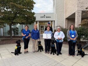 A group of six people, including four women and two men, stand outside a city hall building with three black dogs wearing harnesses. Two women hold leashes and share smiles, with one holding a document. The setting features trees and a paved area.