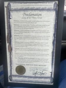 A framed proclamation from the City of El Paso, Texas, declaring September 2025 as 'National Service Dog Month'. The document details the importance of service dogs for individuals with disabilities and highlights the contributions of Canine Companions, a nonprofit organization. It includes a decorative border, a seal of the city, and a signature from the mayor.