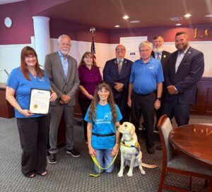 A group photo featuring several people in business attire standing and sitting in a meeting room. In the front, two women are kneeling, one holding a certificate, and the other is accompanied by a yellow Labrador service dog wearing a harness. The background includes a flag and a table.