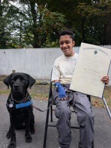 A young person sitting on a chair holds a certificate in one hand while a black service dog sits beside them. They are outdoors, with trees in the background and a white fence behind them.