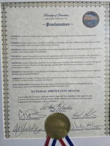 A proclamation from the Township of Hamilton, Atlantic County, NJ, designating September as National Service Dog Month, highlighting the importance of service dogs for people with disabilities, and encouraging community awareness and respect for the rights of those assisted by them. The document features signatures from various officials and a decorative seal.