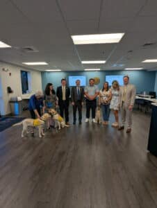 A group of people standing in a bright office setting, featuring two service dogs wearing vests. The group includes a mix of men and women, some in formal attire, smiling and posing for the photo.