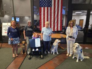 Four women and three dogs pose in front of a large American flag in a community meeting room. One woman is in a wheelchair holding a certificate, while the others stand beside her with two dogs wearing service vests.