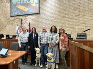 Five people stand together in a courtroom, smiling for the camera. Two yellow Labrador Retrievers, one wearing a blue scarf, sit at their feet. The background features a stone wall with an eagle motif and a large screen displaying a meeting.
