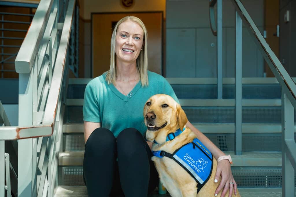 A woman with straight blonde hair sits on a staircase beside a yellow Labrador retriever wearing a blue vest that reads 'Canine Companions'. She is wearing a green top and black pants, smiling at the camera.
