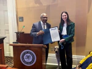 A man in a suit and glasses presents a certificate to a young woman in a green blazer, both smiling. A guide dog sits between them. The background shows a city council setting with a sign indicating 'Council Member'.