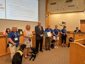A group of six people, including four wearing blue shirts, stands in a meeting room with two service dogs. A man in a suit holds a proclamation. In the background, there’s a presentation screen with text and a panel of officials.
