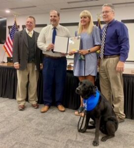 A group of four people stand together in a meeting room, holding a certificate. The individuals include two men in suits, one man in a light-colored shirt and tie, and a woman wearing a blue dress, holding a document and accompanied by a black dog wearing a blue bandana. The room has an American flag in the background.