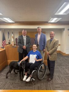 A woman in a blue shirt sits in a wheelchair holding a certificate, accompanied by a black dog. She is surrounded by four men in formal attire standing behind her, with American flags and official decor visible in the background.