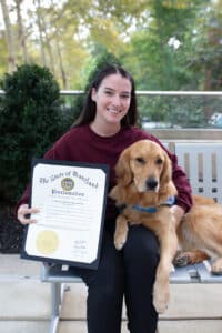 A woman wearing a maroon sweatshirt smiles while sitting on a bench, holding a proclamation from the State of Maryland recognizing National Service Dog Month. A golden retriever sits next to her, resting its head on her lap.
