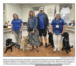 A group of four people stands in front of a desk, each holding or accompanied by a service dog. Two women and a man are wearing blue shirts, while another woman is in a denim jacket. The dogs, one yellow Labrador and two black Labradors, are wearing service vests. In the background, a person is seated at the desk.