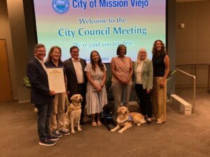 A group of seven people stands together at a City Council meeting in Mission Viejo, with two dogs seated in front; one dog is wearing a yellow harness and the other a blue vest. A large screen in the background displays 'Welcome to the City Council Meeting' and the city seal.