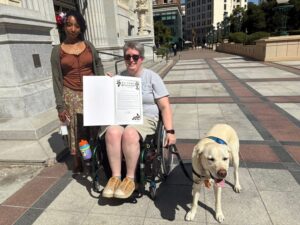 A woman in a wheelchair holds a proclamation document while smiling at the camera. She is wearing a gray shirt and light-colored shorts. Next to her stands another woman with curly hair, wearing a brown top and a patterned skirt. A yellow labrador retriever is beside them, happily panting. The setting appears to be a city plaza with buildings in the background.
