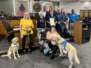 A group photo taken in a city council setting, featuring individuals holding a certificate of recognition, seated and standing around a woman in a wheelchair accompanied by service dogs. The backdrop includes an American flag and city emblem.