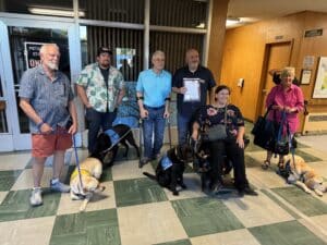 A group of six people, including two seated in wheelchairs, stand indoors next to their guide dogs. The background features large windows and a wooden wall with a city government sign. One man holds a certificate, while another wears a blue short-sleeve shirt. Dogs are wearing vests indicating their training, with one lying down on the floor.