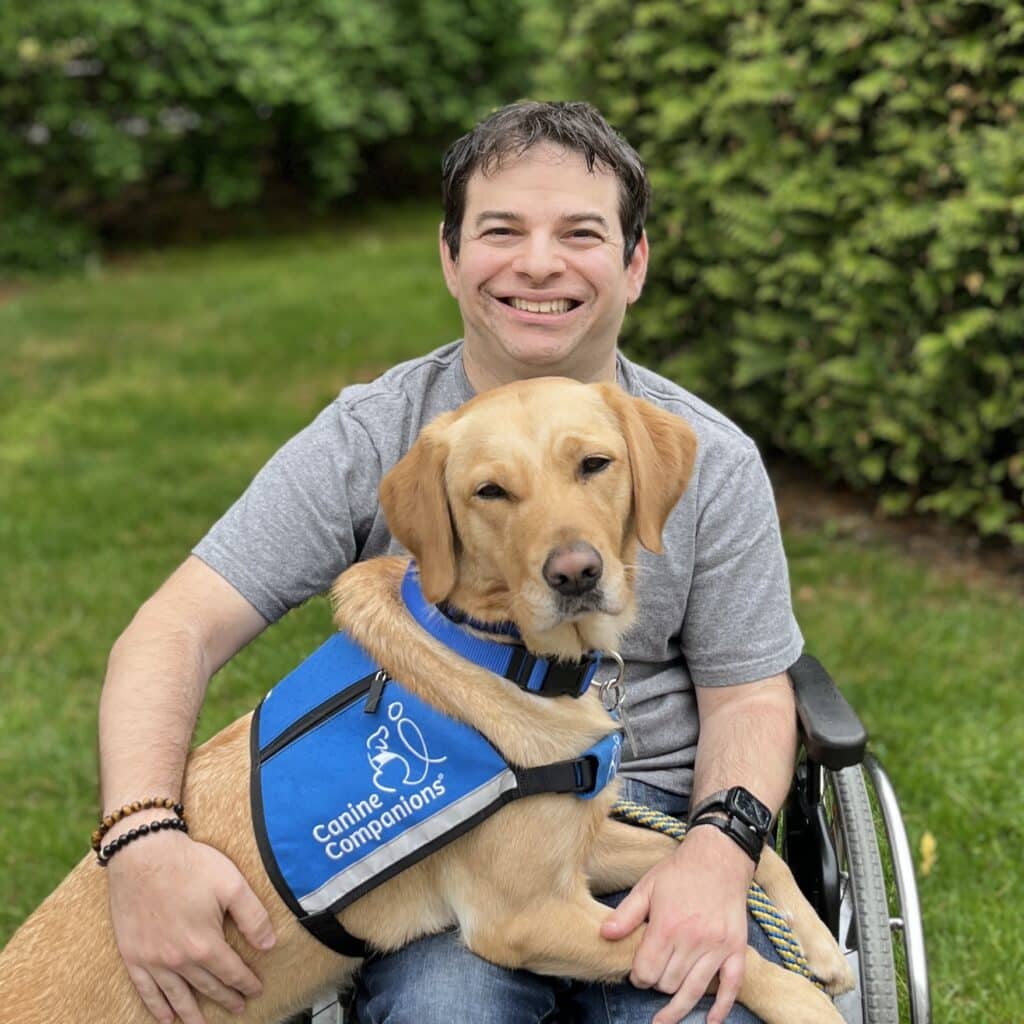 A smiling man in a wheelchair is sitting on grass, holding a golden retriever wearing a blue vest that reads 'Canine Companions'. The background features lush green bushes.