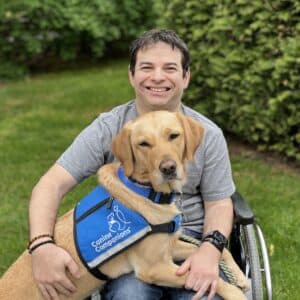 A smiling man in a wheelchair is sitting on grass, holding a golden retriever wearing a blue vest that reads 'Canine Companions'. The background features lush green bushes.