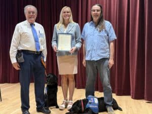 Three individuals stand on stage in front of a red curtain, with two black service dogs beside them. The woman in the center holds a certificate, wearing a light blue blazer and a cream-colored skirt. To her left is an older man in a white shirt and blue tie, while to her right is a man in a blue shirt with long hair. The dogs are wearing service dog vests.