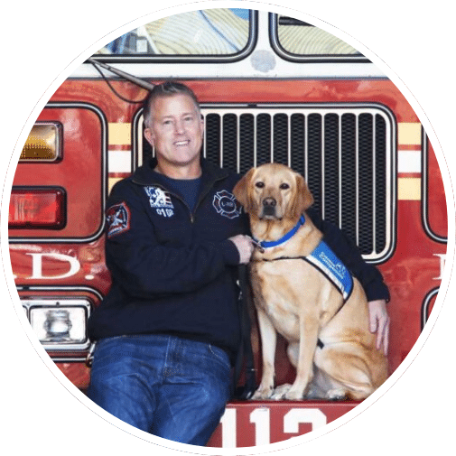 A firefighter posing with a yellow Labrador retriever in front of a fire truck, showcasing a friendly and supportive relationship between them.