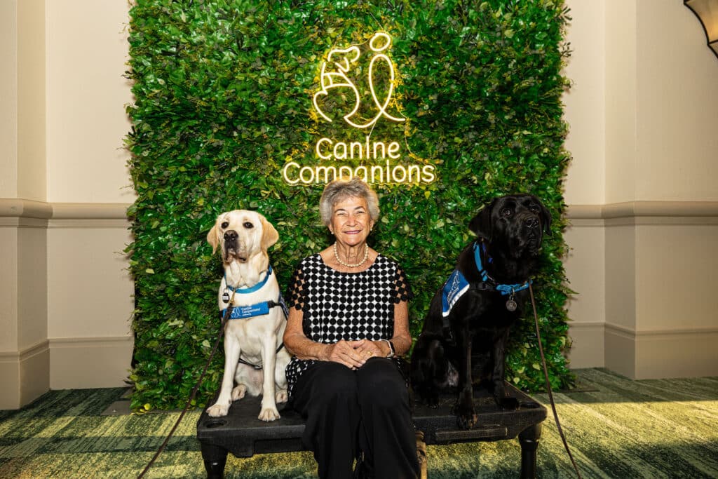 An older woman sits between two trained service dogs, a yellow Labrador and a black Labrador, in front of a green foliage backdrop with a glowing sign that reads 'Canine Companions'. The woman has short gray hair and wears a black and white polka dot shirt.