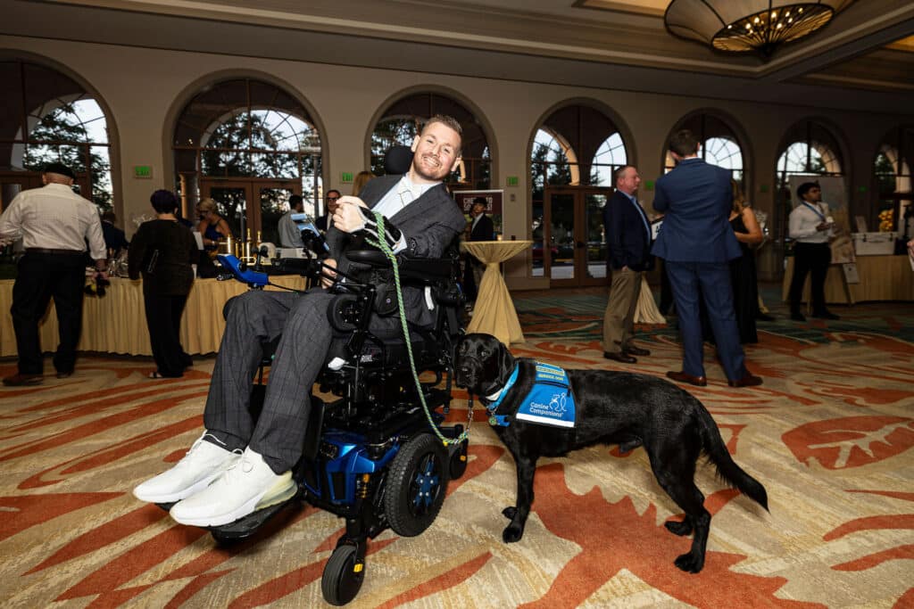 A man in a wheelchair smiles while holding a leash attached to a black service dog wearing a blue vest labeled "Canine Companions." They are in a large room with informational displays and other guests mingling in the background.